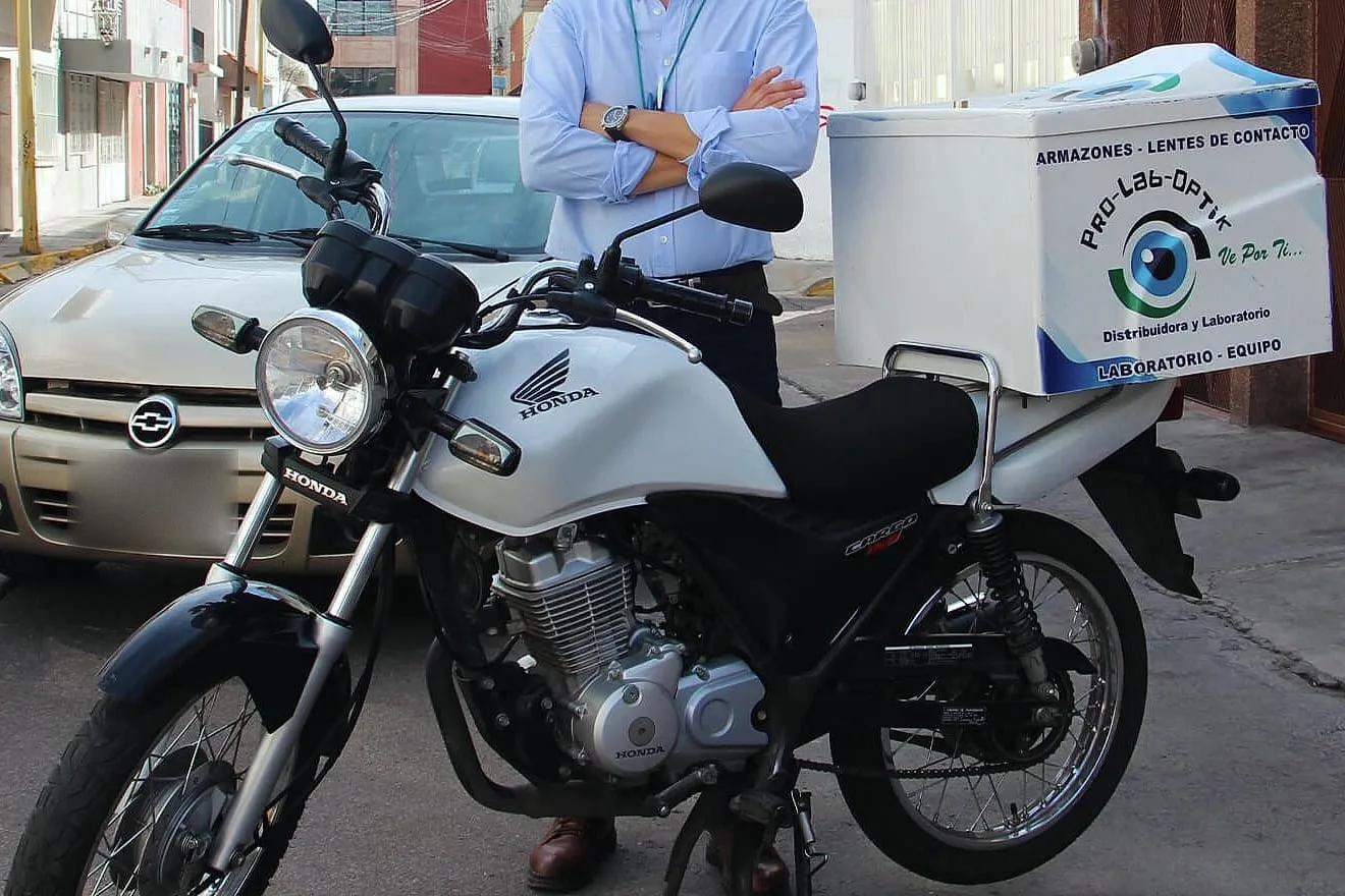 Motocicleta Honda blanca con baúl de reparto rotulado con la marca del laboratorio; detrás, una persona con camisa azul y brazos cruzados