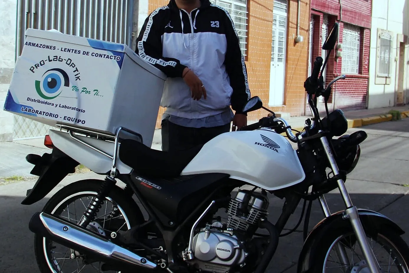 Persona con chamarra deportiva negra y blanca junto a una motocicleta con baúl de reparto rotulado con la marca del laboratorio