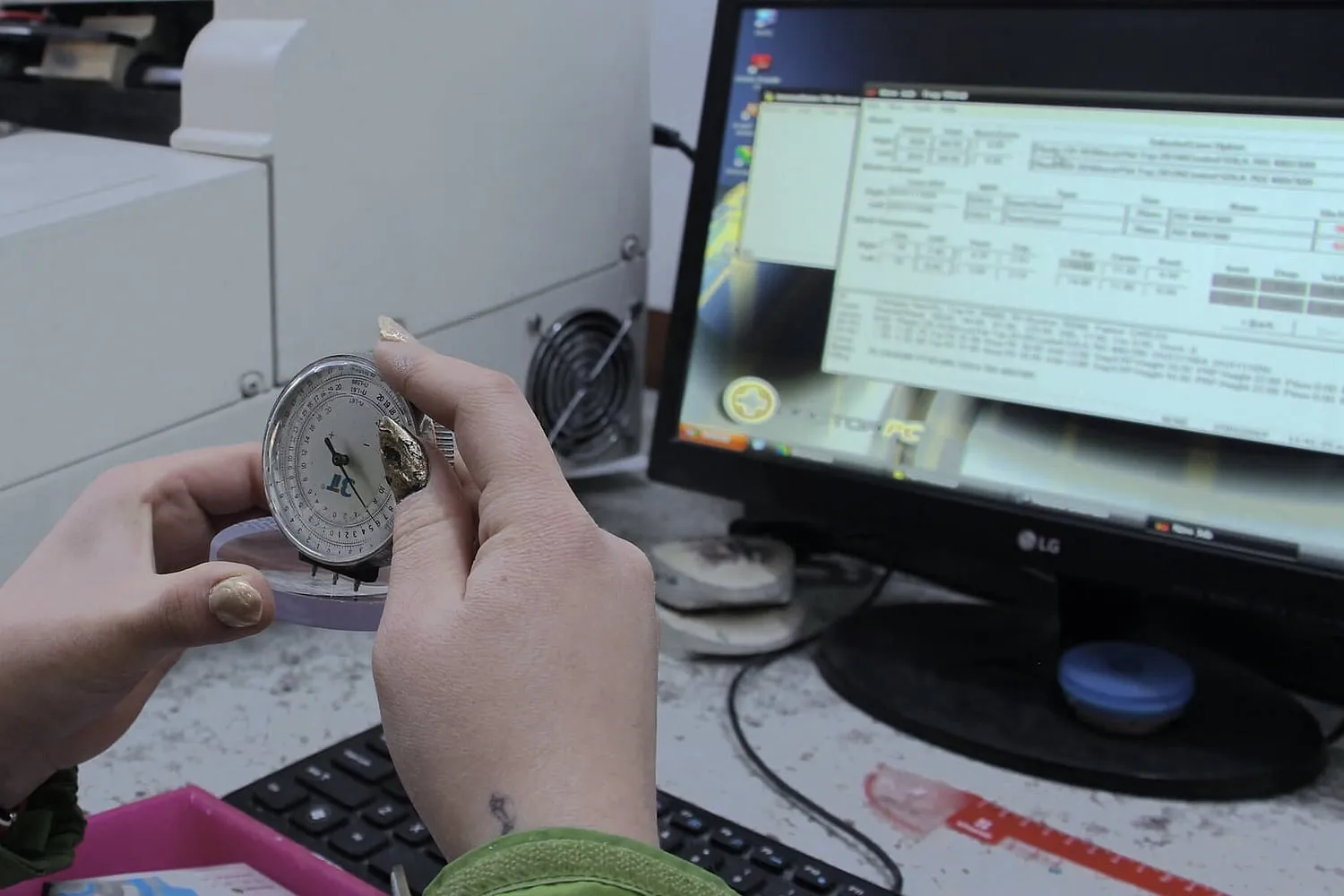 Manos sosteniendo un medidor de lentes sobre una lente, con una computadora con software de tallado al fondo