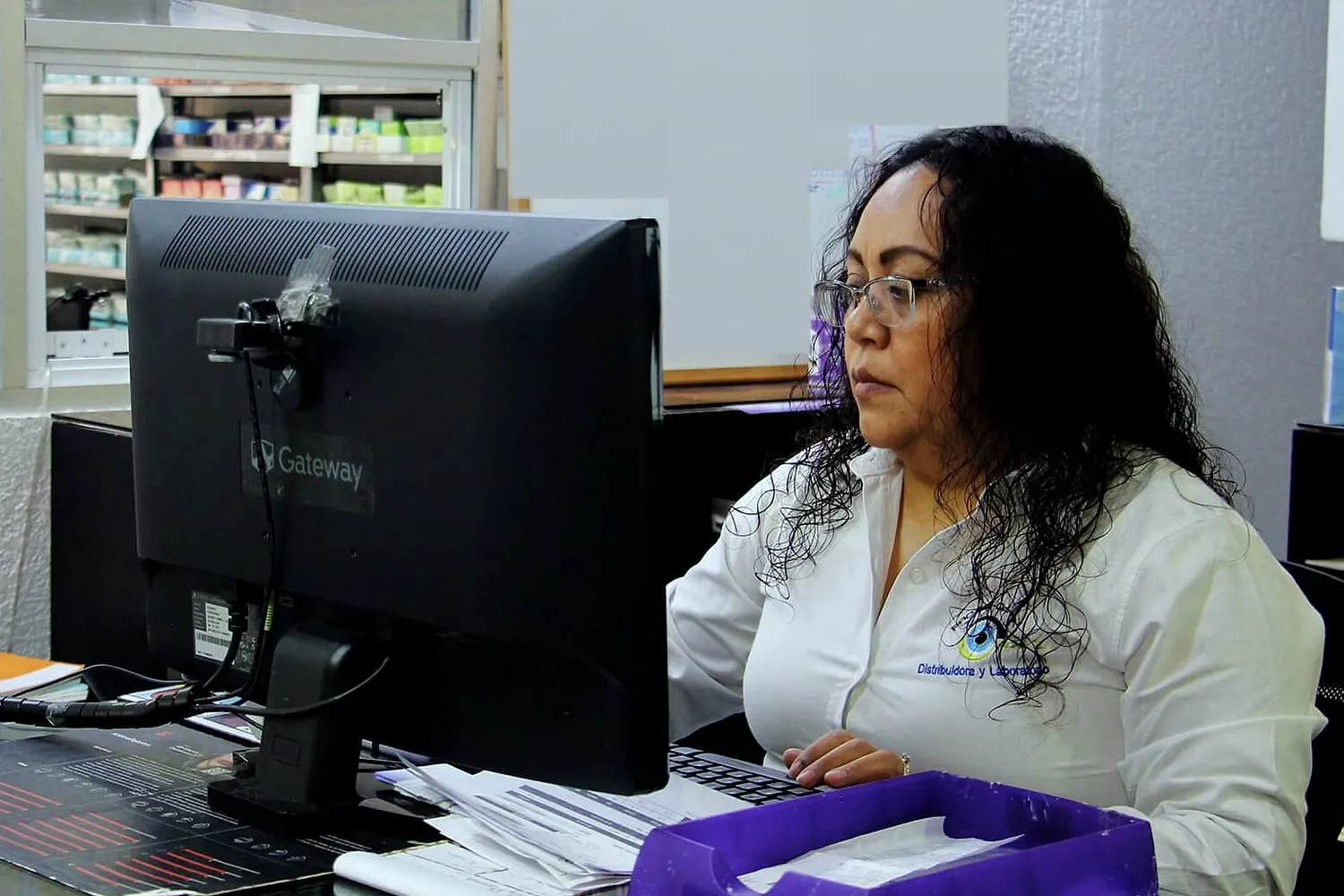 Colaboradora de perfil trabajando en la computadora, con una estantería de cajas de lentes al fondo