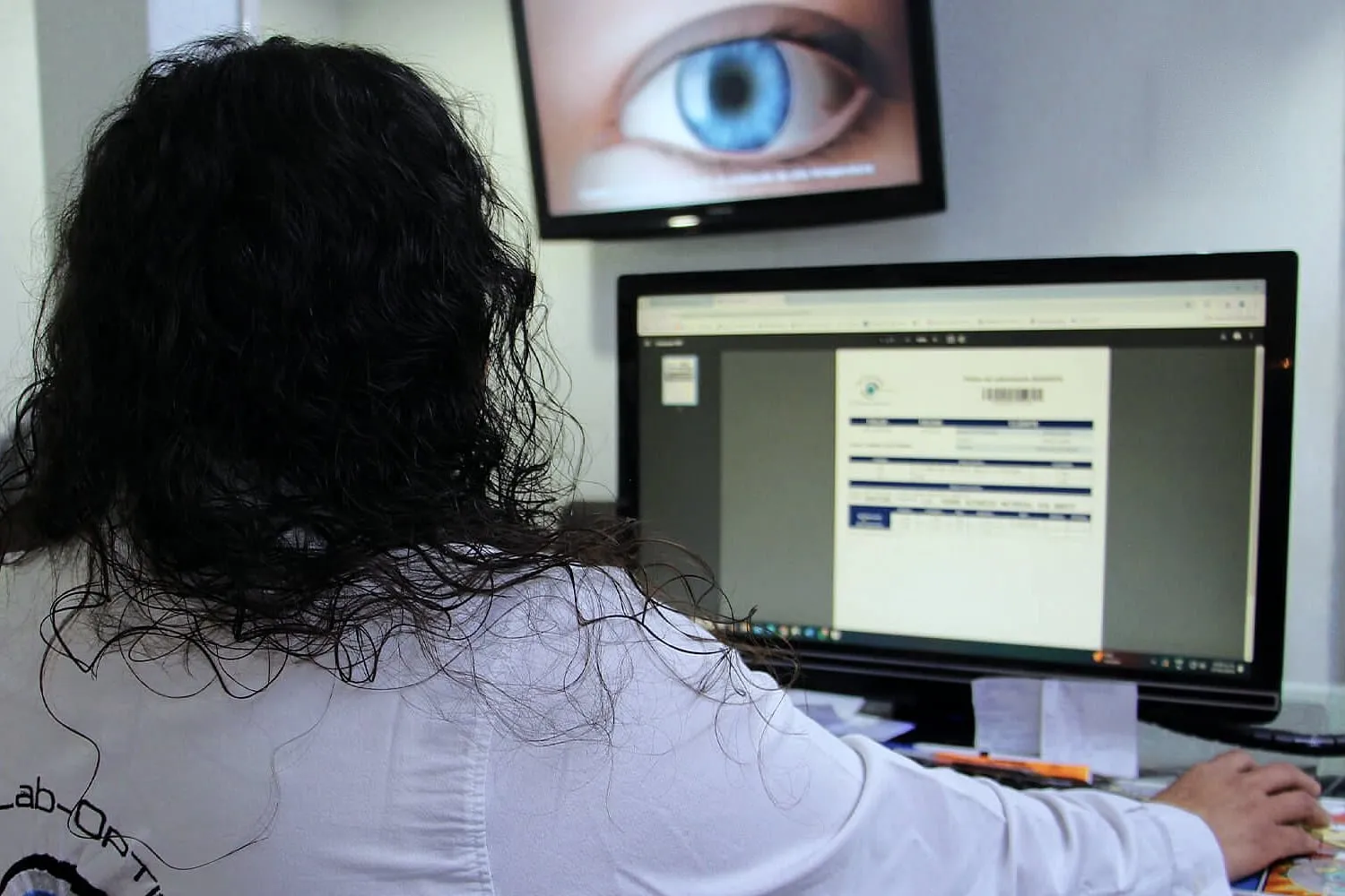 Colaboradora de espaldas, con bata del laboratorio, frente a una computadora; arriba, un televisor con la imagen de un ojo